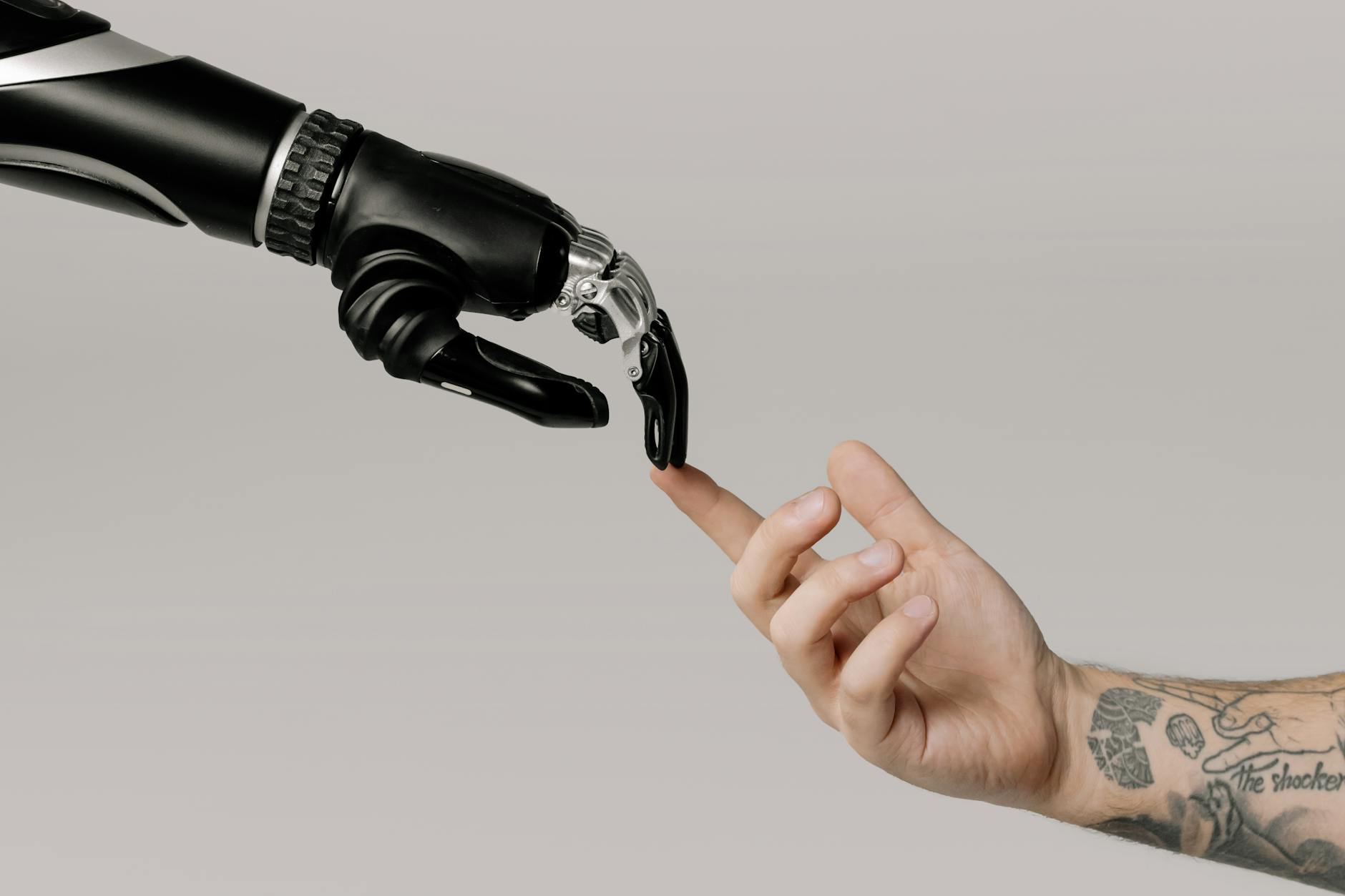 A human hand with tattoos reaching out to a robotic hand on a white background.